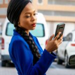 Woman in blue coat with braided hair using smartphone on a city street.